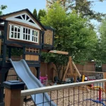 A backyard play structure featuring a slide and two swings, surrounded by grass and trees.