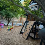 A play area featuring a swing set beside a large tree, inviting children to play and enjoy the outdoors.